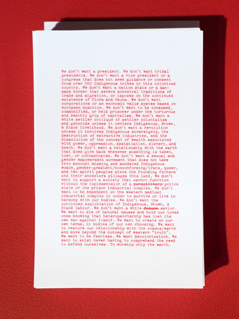 A long text printed in red ink on white paper and affixed to a red gallery wall
