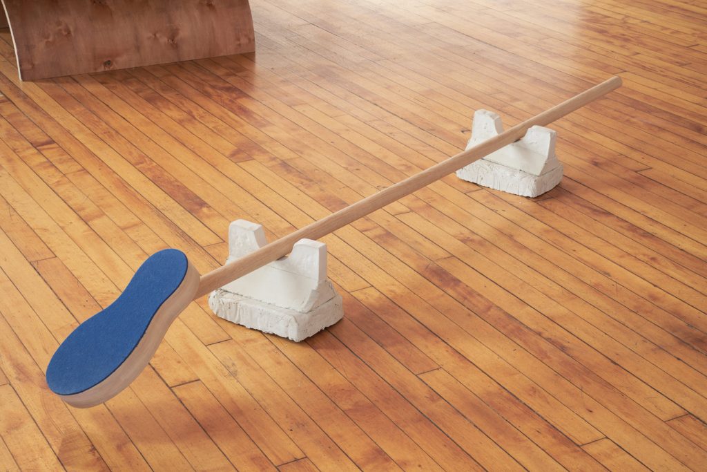 A sculpture comprised of two structures on the floor holding a stick with the sole of a shoe at the end of it.