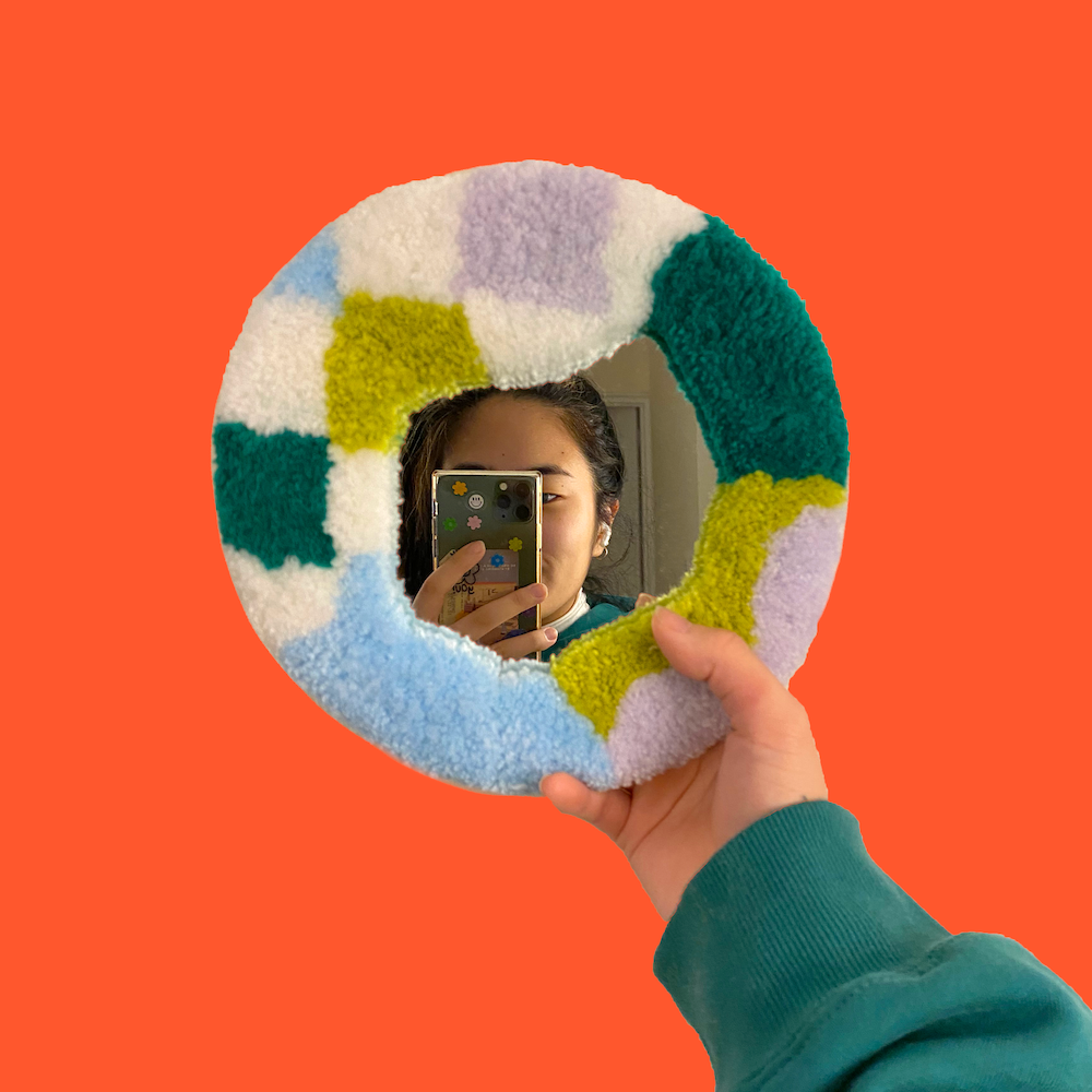 A woman holds a circular mirror with a frame made from atufted rug pattern